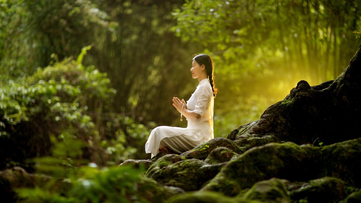 A woman meditating outdoors with prayer beads amidst lush forest greenery.